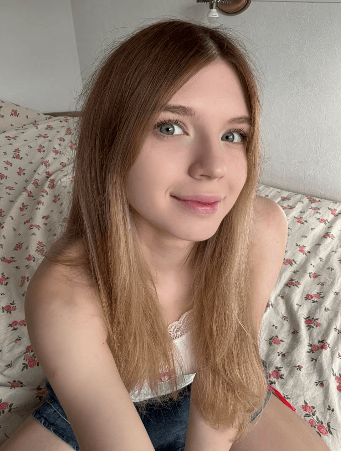 Young woman with long light brown hair sits on a floral bedspread, smiling at the camera.