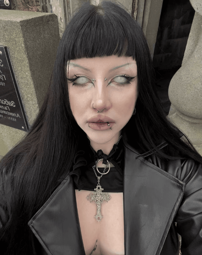 Person with blunt-black hair and gothic makeup wearing a black leather jacket and a large ornate cross pendant on the chest plate necklace, looking upward.