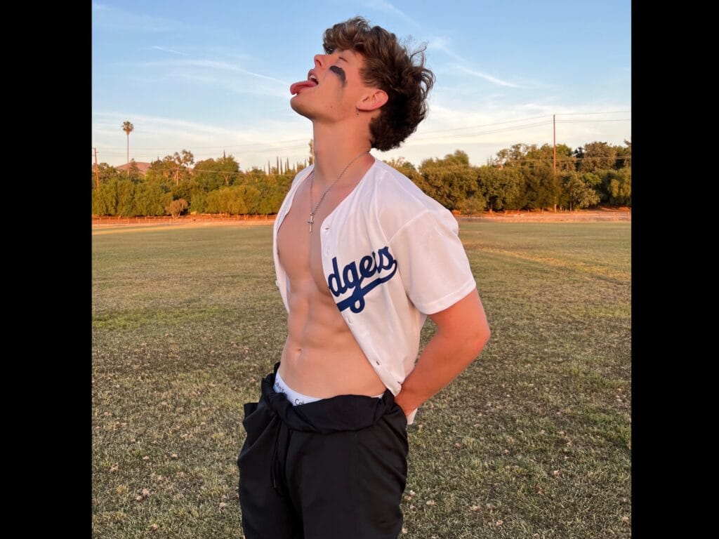 Young man outdoors in a park, unbuttoned Dodgers shirt, tongue sticking out, head tilted back at sunset.