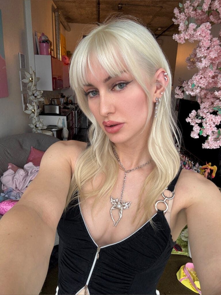 Selfie of a blonde woman in a black zip-front top, wearing silver jewelry, with pink blossoms in the background.