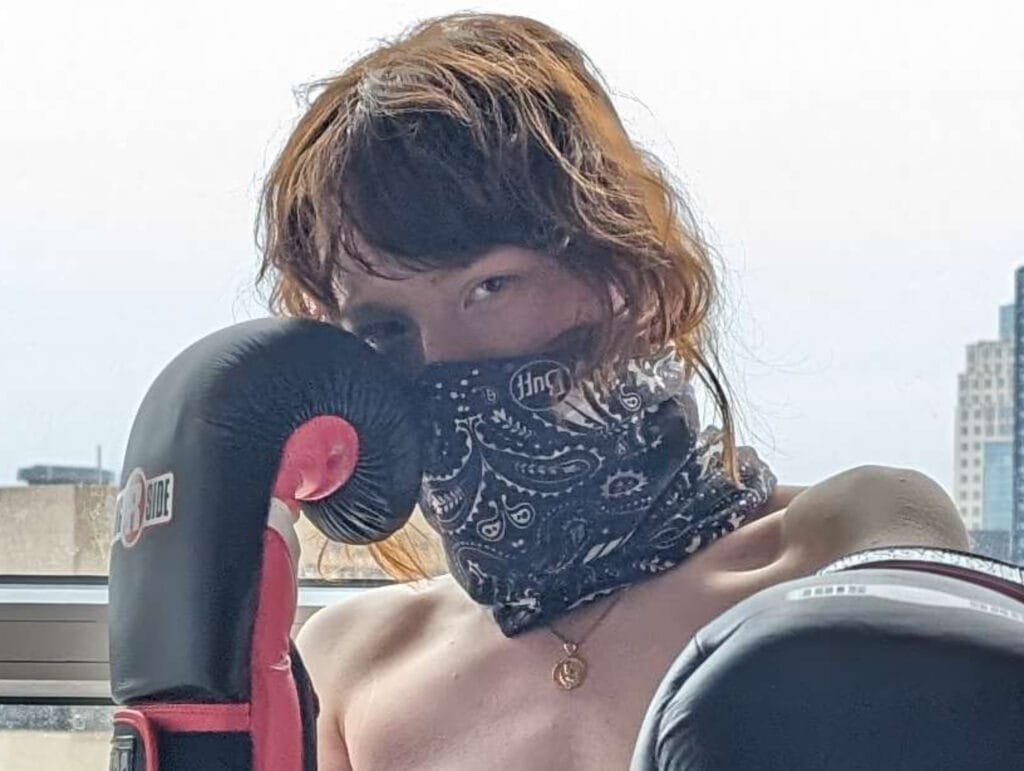 Person outdoors with a patterned scarf over the lower face, wearing a black boxing glove on the left hand, city buildings in the background.