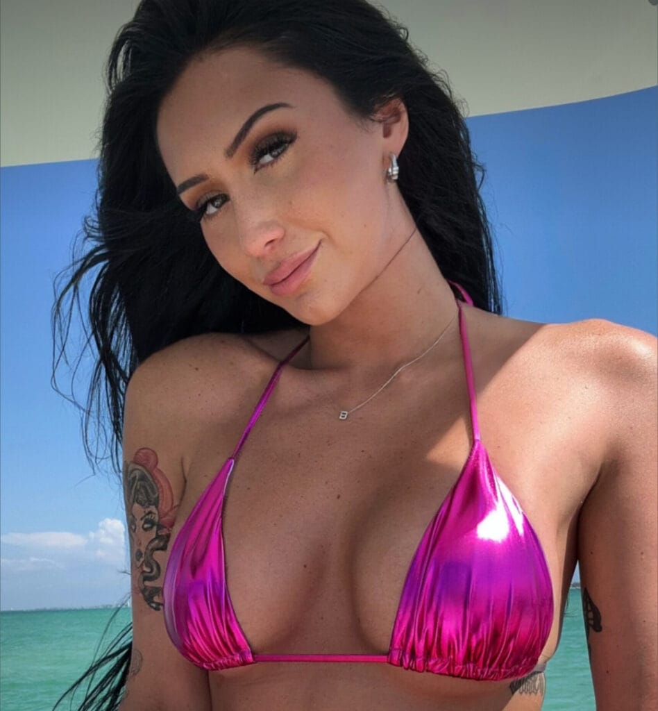 Woman in a pink bikini top at the beach with turquoise water in the background; tattoo on her left arm and a small necklace visible.