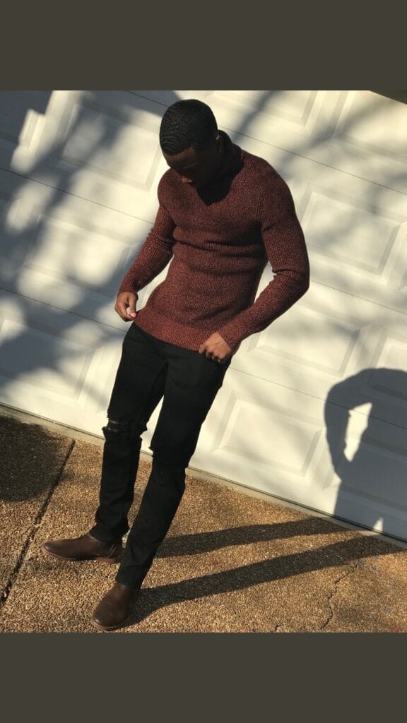 Man in a maroon knit sweater, black ripped jeans and brown boots standing in front of a light garage door outside.