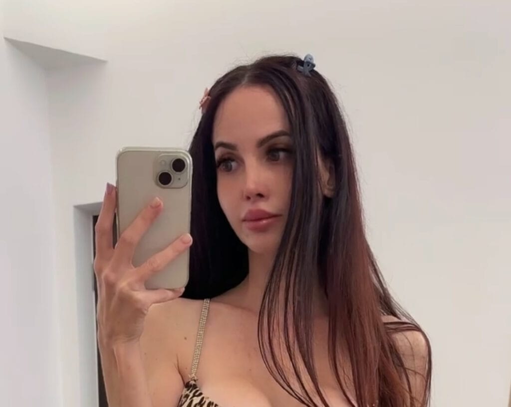 Woman taking a mirror selfie with a gold iPhone, wearing a leopard-print top and hair clips in a bright room.